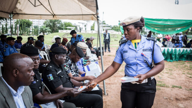 A police officer at an outdoor event passing out pamphlets to an audience of seated officers