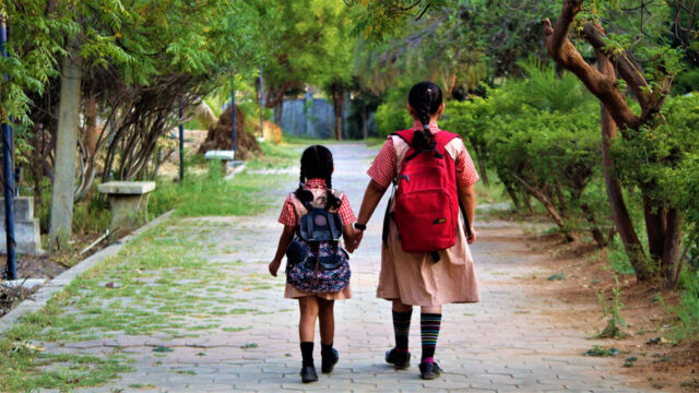 Two schoolgirls with backpacks holding hands while walking through a town path