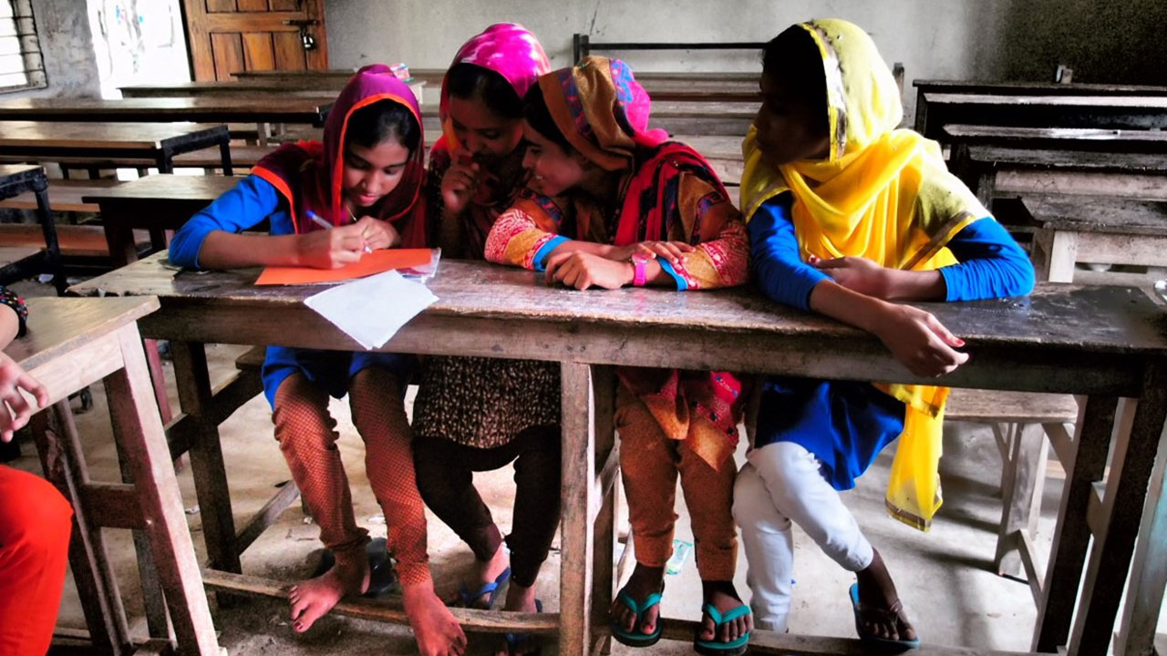Four schoolgirls sitting at a desk while with one writing on a piece of paper