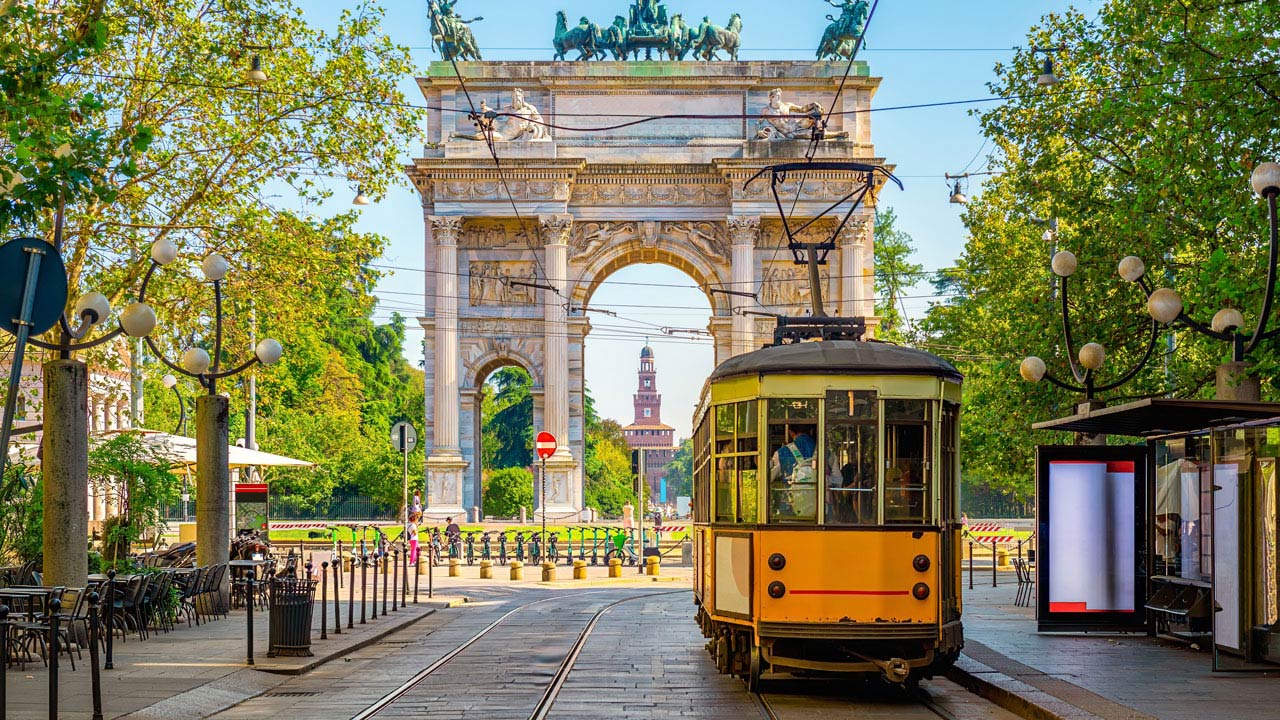 A yellow tram passing under a grand archway, surrounded by trees and outdoor seating in a vibrant city setting