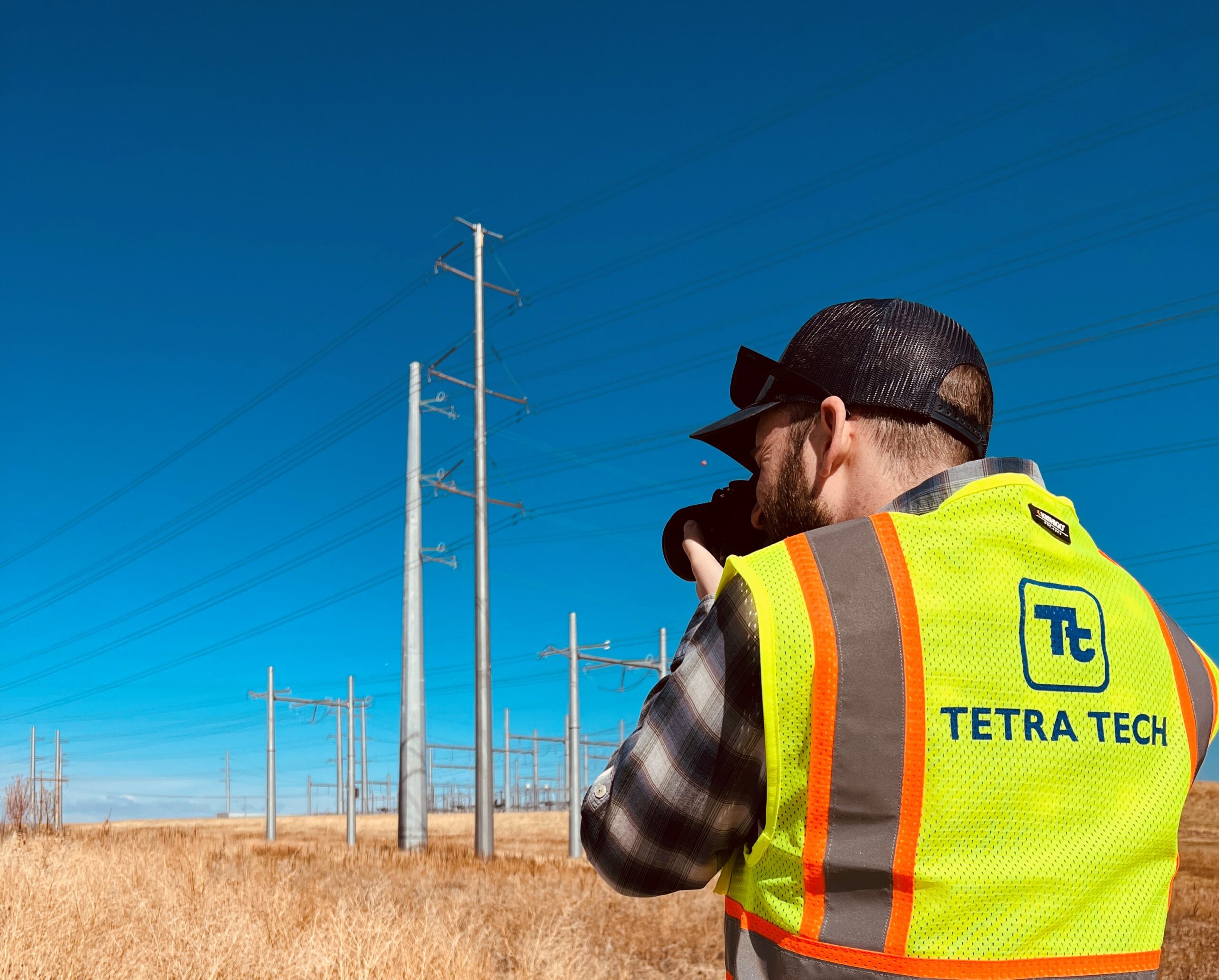 A man is taking a technical photograph of electricity infrastructure. He is wearing a yellow hi-vis vest branded with the logo of Tetra Tech.