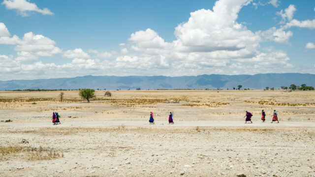 A group of people in colorful attire walking across a dry landscape under a bright blue sky with fluffy clouds