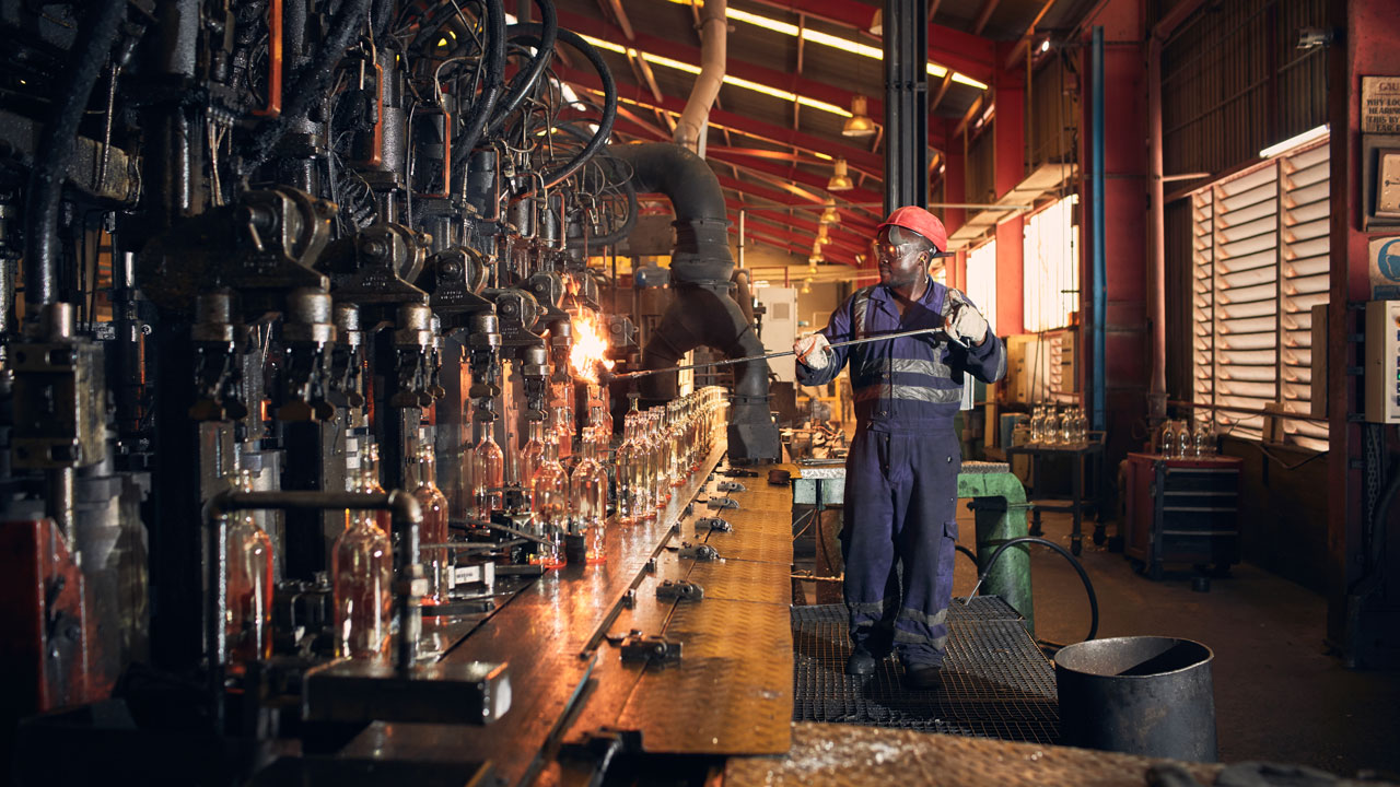 A factory worker in protective gear using a metal rod to work with hot glass on a conveyor belt of glass bottles