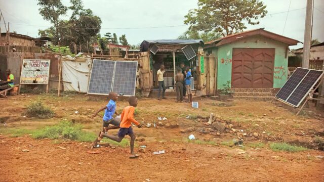 Children frolicking towards a solar panel by tin shack in a Sub-Saharan community