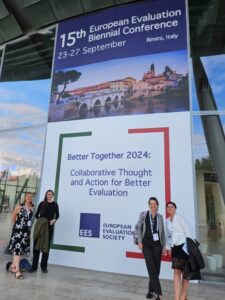 Four team members standing in front of the advertising for the European Evaluation Society Conference in Rimini, Italy, September 2024