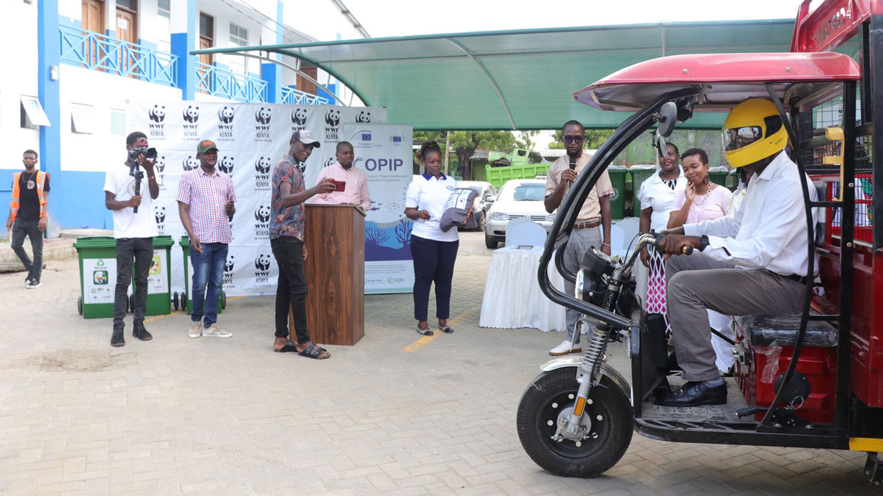 Waste collection vehicle driver with a helmet driving a tuk-tuk next to a COPIP event tent