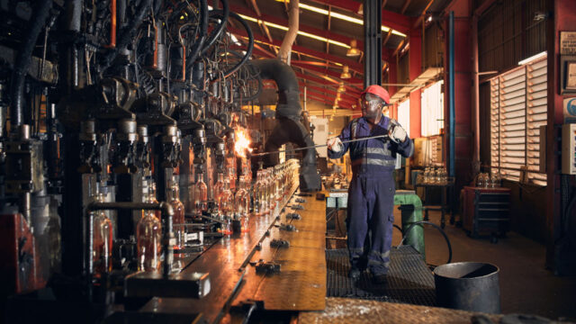 A factory worker in protective gear using a metal rod to work with hot glass on a conveyor belt of glass bottles