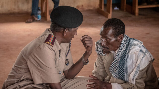 An officer of a Kenya Administration police unit in beige uniform squatting next to and talking to a civilian