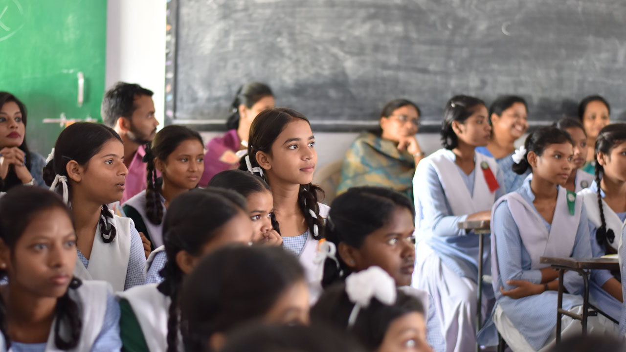 A group of students in uniforms attentively listen in a classroom, with a blackboard visible in the background