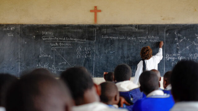 A teacher writing on a chalkboard in a classroom with students watching from their seats