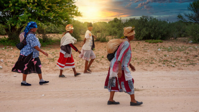 Four women carrying bags hiking down a dirt road