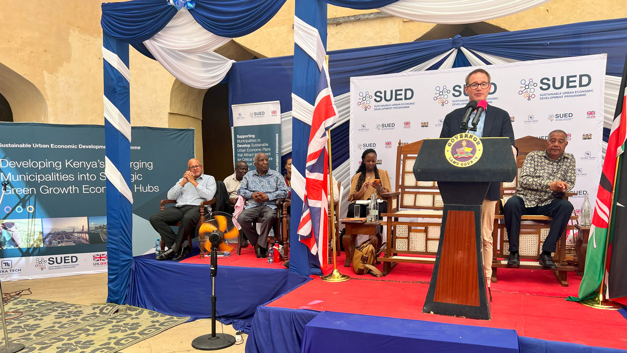 A speaker standing at a podium with attendees seated nearby during an event for SUED Kenya