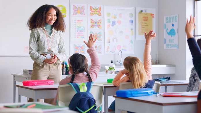 Female teacher in classroom with pupils raising their hands.