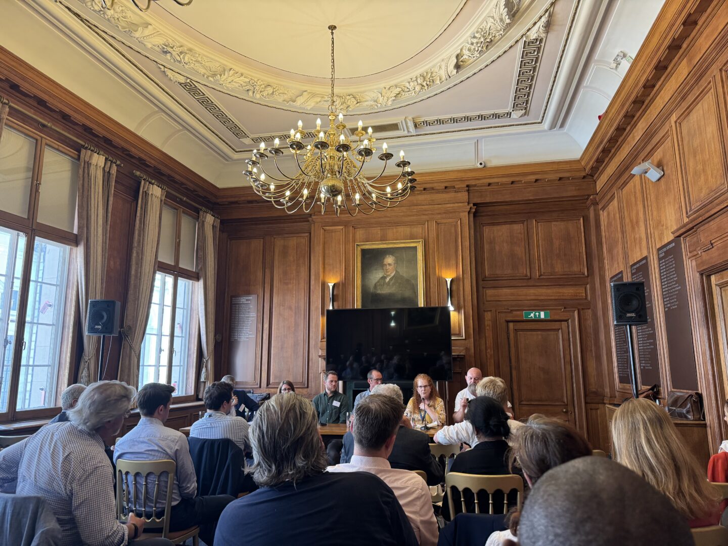 People listening to a panel discussion comprising five panellist, in an classical style room with wood panelling and large windows to the left.